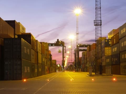 Stacked shipping containers at a sea freight terminal at dusk with port cranes in the background