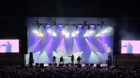 Outdoor festival stage at night with band performing, ground-stacked PA speaker systems flanking the stage, LED screens and purple beam lighting